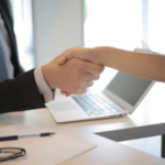 Two professionals shaking hands over a desk with an open laptop, glasses, and a pen indicating a business agreement or greeting.