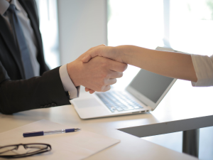 Two professionals shaking hands over a desk with an open laptop, glasses, and a pen indicating a business agreement or greeting.