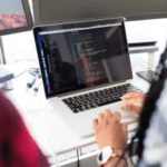 Person working on a laptop with code on the screen, sitting at a desk with a mountain view outside the window.