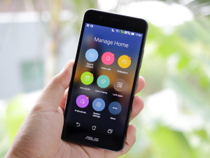 Hand holding an ASUS smartphone with a smart home management app open, displaying colorful icons on the screen, with a blurred green foliage background.