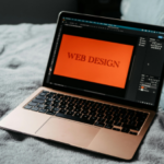 Laptop on a grey bedspread displaying WEB DESIGN on screen with graphic design software active.