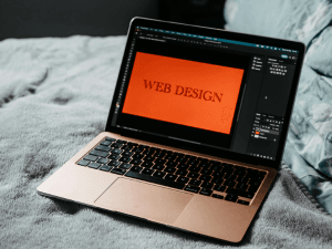 Laptop on a grey bedspread displaying WEB DESIGN on screen with graphic design software active.