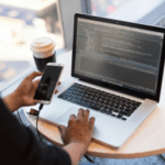 Person using a laptop with code on the screen while holding a smartphone, with a coffee cup and glasses on the table.