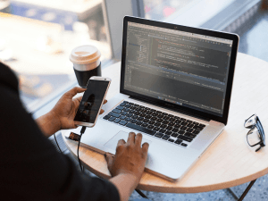 Person using a laptop with code on the screen while holding a smartphone, with a coffee cup and glasses on the table.