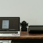 A laptop with stickers on it opened to a design program, a decorative statue, and a printer on a wooden desk against a plain wall.