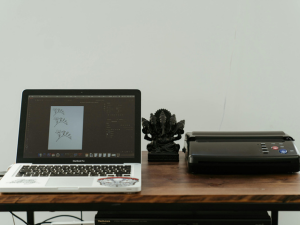 A laptop with stickers on it opened to a design program, a decorative statue, and a printer on a wooden desk against a plain wall.