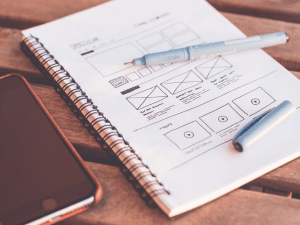 A close-up of a web design wireframe sketch on a spiral notebook, with a blue pen on top, beside a smartphone on a wooden surface.