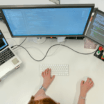 A top-down view of a workspace with a person typing on a keyboard, a laptop, and a monitor displaying code, surrounded by desk accessories.