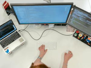 A top-down view of a workspace with a person typing on a keyboard, a laptop, and a monitor displaying code, surrounded by desk accessories.