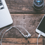 Laptop, smartphone with earphones, and camera lens on wooden surface.