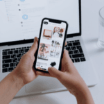 Person holding a smartphone with Pinterest on the screen in front of an open laptop on a white desk.