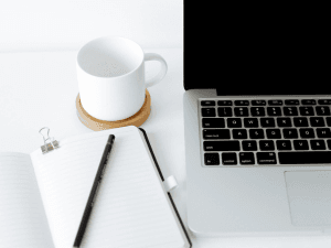 A clean and simple workspace featuring an open notebook with a pen on top, a white mug on a coaster, and a laptop with black keys on a white desk.