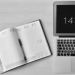 Black and white photo of an open planner and a pen next to a laptop displaying the time 14:41 on a wooden desk.