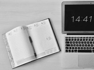 Black and white photo of an open planner and a pen next to a laptop displaying the time 14:41 on a wooden desk.