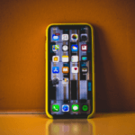 Smartphone with colorful icons on screen, encased in a yellow bumper, standing upright on a reflective surface against an orange background.