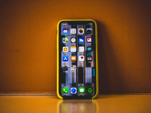 Smartphone with colorful icons on screen, encased in a yellow bumper, standing upright on a reflective surface against an orange background.