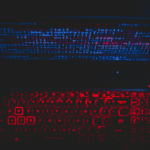 Alt text: Backlit keyboard with red illumination against a dark background, with the glow of coding text on a computer screen above.