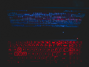Alt text: Backlit keyboard with red illumination against a dark background, with the glow of coding text on a computer screen above.