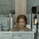 Alt text: A focused woman wearing a headset working at a computer desk in a modern office environment with partitions and colleagues in the background.
