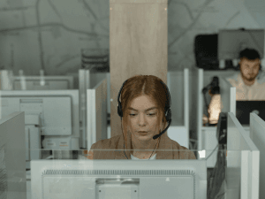 Alt text: A focused woman wearing a headset working at a computer desk in a modern office environment with partitions and colleagues in the background.