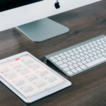 An Apple iMac with keyboard on a wooden desk, alongside an iPad displaying a calendar.