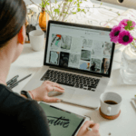 Over-the-shoulder view of a woman working on a laptop with mood board images on the screen, sitting at a desk with a notepad, cup of coffee, and vase of pink flowers.