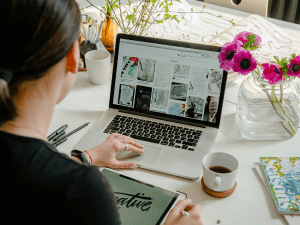 Over-the-shoulder view of a woman working on a laptop with mood board images on the screen, sitting at a desk with a notepad, cup of coffee, and vase of pink flowers.