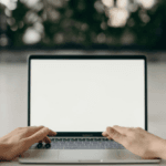 Person typing on a laptop with a blank white screen, set on a table with a blurred background of indoor plants.