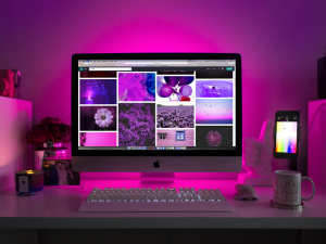 A desktop workspace illuminated by purple ambient lighting featuring an iMac with a photo collage on the screen, a smartphone, a keyboard, a mouse, a mug, and decorative plants.