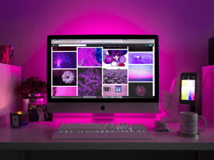 A desktop workspace illuminated by purple ambient lighting featuring an iMac with a photo collage on the screen, a smartphone, a keyboard, a mouse, a mug, and decorative plants.