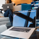 An open laptop with code on the screen in focus on a glass table with a blurred person working in the background.