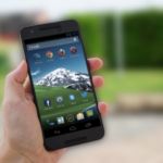 Hand holding a smartphone with a mountain wallpaper on screen, blurred background of a residential area.