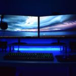 A dimly lit home office setup with two large monitors displaying a dramatic sky wallpaper, an illuminated keyboard with green backlight, a mouse, and under-desk LED lighting casting a blue glow.