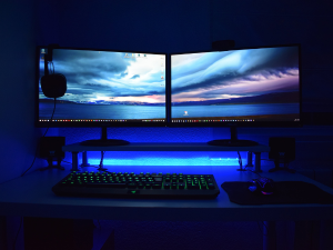 A dimly lit home office setup with two large monitors displaying a dramatic sky wallpaper, an illuminated keyboard with green backlight, a mouse, and under-desk LED lighting casting a blue glow.