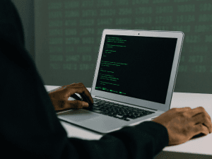 Alt text: A person coding on a laptop with a screen displaying lines of code, set against a dark backdrop with out-of-focus digital data.