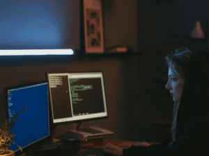 A person working intently on a computer with multiple screens in a dark room, illuminated by the soft glow of the monitors.