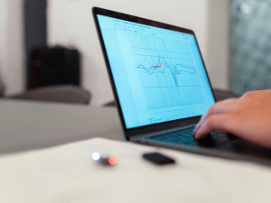 Close-up of a person's hand using a laptop displaying graphs, with a smartphone and wireless earbuds on the table.